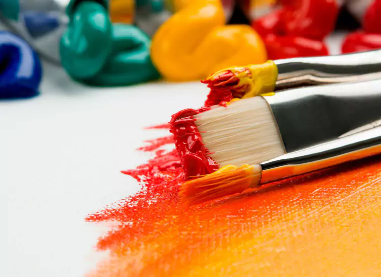 A brush with paint red and yellow paint in the foreground laying on a canvas and dollops of paint in the background. Photo by Anna Kolosyuk Unsplash.